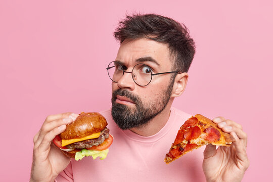 Eating junk food. Serious attentive bearded adult European man holds tasty hamburger and slice of pizza wears spectacles eats cheat meal isolated over pink background feels hungry after work