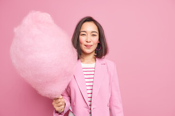 Pretty young Asian woman with dark hair holds tasty cotton candy has sweet tooth dressed in stylish clothes being in good mood isolated over pink background enjoys spare time during weekend.