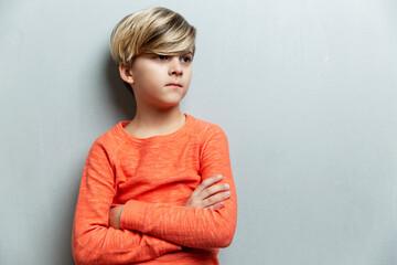 A serious boy of 9 years old in an orange jacket stands against the wall. The arms are crossed over the chest. Gray background. © Анна Демидова