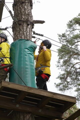 Children scouts in helmets stand on a high-rise base platform on a tree on European outdoor forest rope park at spring summer day - climbing sports active recreation
