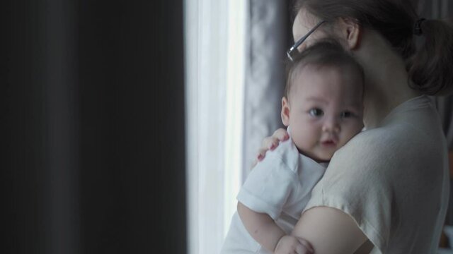 portrait of happy asian woman mother holding sleepy baby infant near window at home daytime. 4 months baby embracing in parent arm. relationship mother and daughter. mom touching baby lull to sleep.