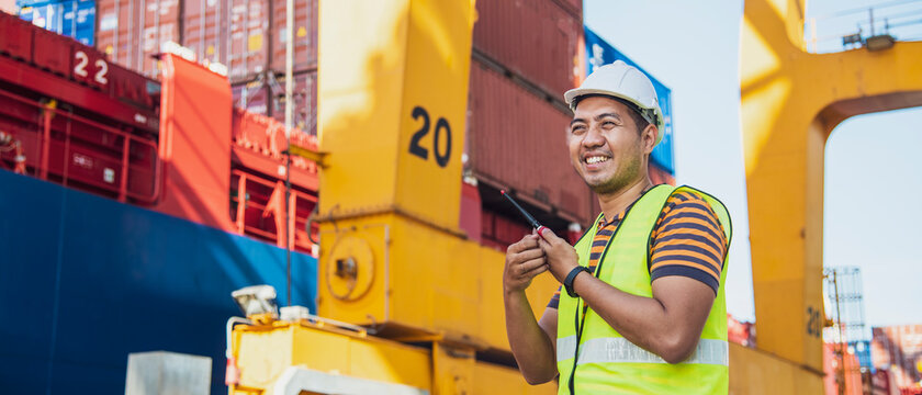 portrait photo of happy Asian logistics worker with radio transceiver working in port shipping containers. logistics, sea transportation,  commercial and logistics business concept