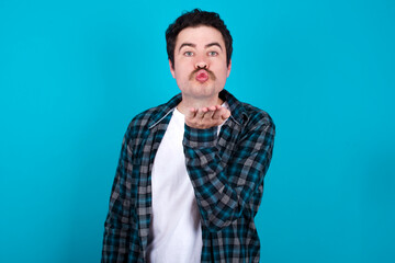young Caucasian man with moustache wearing plaid shirt against blue wall looking at the camera blowing a kiss with hand on air being lovely and sexy. Love expression.