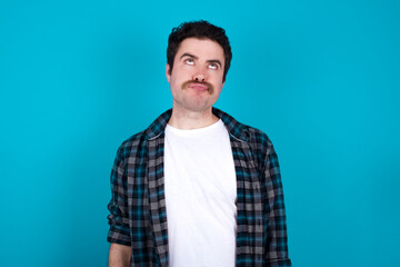 Fototapeta premium young Caucasian man with moustache wearing plaid shirt against blue wall has worried face looking up lips together, being upset thinking about something important, keeps hands down.