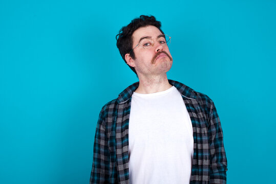 young Caucasian man with moustache wearing plaid shirt against blue wall with snobbish expression curving lips and raising eyebrows, looking with doubtful and skeptical expression, suspect and doubt.