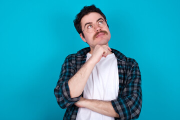 Face expressions and emotions. Thoughtful young Caucasian man with moustache wearing plaid shirt against blue wall holding hand under his head, having doubtful look.