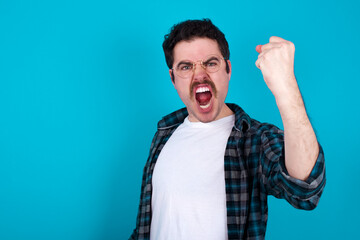 Fierce young Caucasian man with moustache wearing plaid shirt against blue wall holding fist in front as if is ready for fight or challenge, screaming and having aggressive expression on face.