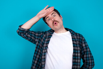 young Caucasian man with moustache wearing plaid shirt against blue, wiping forehead with hand...