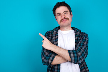 young Caucasian man with moustache wearing plaid shirt against blue wall smiling broadly at camera, pointing fingers away, showing something interesting and exciting.