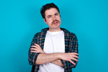 young Caucasian man with moustache wearing plaid shirt against blue wall frowning his face in...