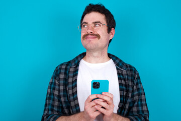 Happy young Caucasian man with moustache wearing plaid shirt against blue wall listening to music with earphones using mobile phone.