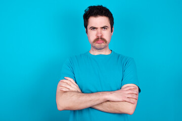 Gloomy dissatisfied young handsome Caucasian man with moustache wearing blue t-shirt against blue background looks with miserable expression at camera from under forehead, makes unhappy grimace