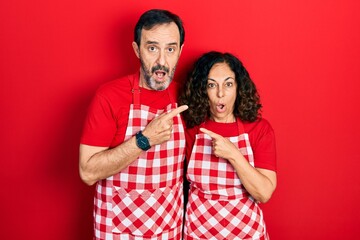 Middle age couple of hispanic woman and man wearing cook apron surprised pointing with finger to the side, open mouth amazed expression.