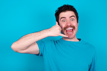 young handsome Caucasian man with moustache wearing blue t-shirt against blue background makes phone gesture, says call me back again, has glad expression.