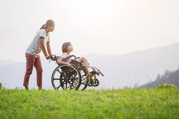 Obraz premium Disabled handicapped child sitting in wheelchair and care helper walking on mountain meadow park in sunny day.. International Disability Day.