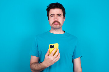 Upset dissatisfied young handsome Caucasian man with moustache wearing blue t-shirt against blue wall uses mobile software application and surrfs information in internet, holds modern mobile hand