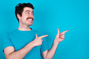 Optimistic young handsome Caucasian man with moustache wearing blue t-shirt against blue background points with both hands and  looking at empty space.