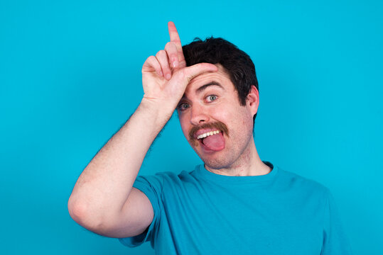 Funny young handsome Caucasian man with moustache wearing blue t-shirt against blue background makes loser gesture mocking at someone sticks out tongue making grimace face.