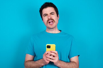 Photo of astonished crazy young handsome Caucasian man with moustache wearing blue t-shirt against blue background hold smartphone dislike feedback concept