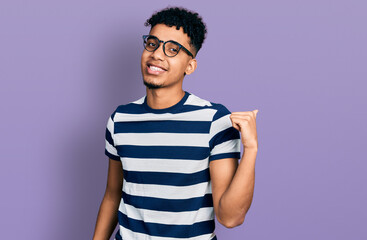 Young african american man wearing casual clothes and glasses smiling with happy face looking and pointing to the side with thumb up.