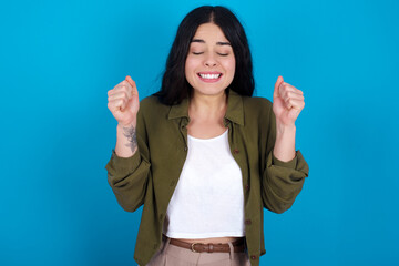 young beautiful tattooed girl standing against blue background rejoicing his success and victory clenching fists with joy being happy to achieve aim and goals. Positive emotions, feelings.