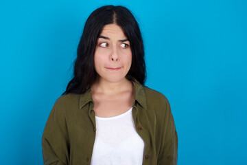 Fototapeta premium Amazed puzzled young beautiful tattooed girl standing against blue background, curves lips and has worried look, sees something awful in front.