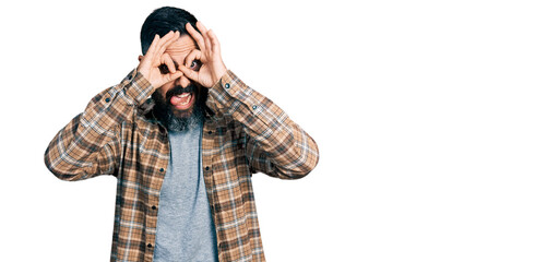 Hispanic man with beard wearing casual shirt doing ok gesture like binoculars sticking tongue out, eyes looking through fingers. crazy expression.