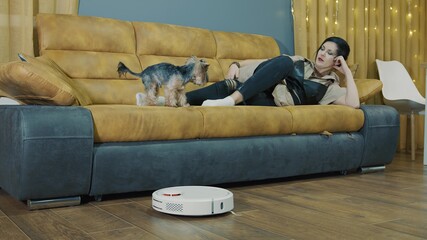 Woman resting at home on the couch and a robot vacuum cleaner vacuuming the room. While the robot is running, the woman is resting. The future in cleaning the room.