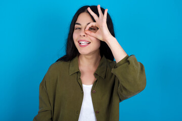 young beautiful tattooed girl standing against blue wall with happy face smiling doing ok sign with hand on eye looking through finger.