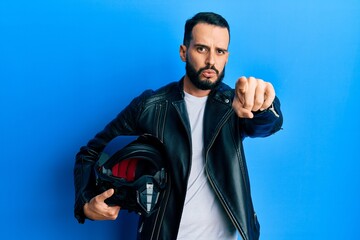Young man with beard holding motorcycle helmet pointing with finger to the camera and to you, confident gesture looking serious