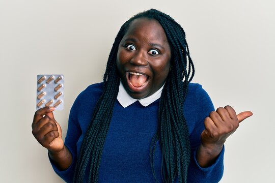 Young black woman with braids holding pills pointing thumb up to the side smiling happy with open mouth