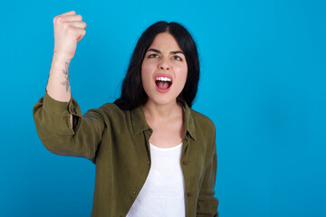 Fierce young beautiful tattooed girl standing against blue background holding fist in front as if is ready for fight or challenge, screaming and having aggressive expression on face.