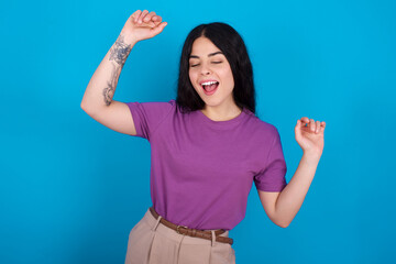Fototapeta premium Photo of upbeat young beautiful tattooed girl wearing purple t-shirt standing against blue has fun and dances carefree wear being in perfect mood makes movements. Spends free time on disco party