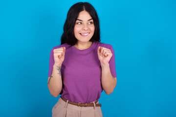 young beautiful tattooed girl wearing purple t-shirt standing against blue background clenches fists and awaits for something nice happened looks away bites lips and waits announcement of results