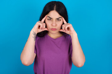 Serious concentrated young beautiful tattooed girl wearing blue t-shirt standing  keeps fingers on temples, tries to ease tension, gather with thoughts and remember important information for exam