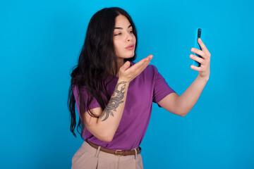 young beautiful tattooed girl wearing blue t-shirt standing against blue background blows air kiss at camera of smartphone and takes selfie, sends mwah via online call.