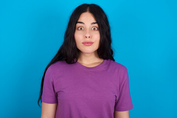 Fototapeta premium Stunned young beautiful tattooed girl wearing blue t-shirt standing against blue background stares reacts on shocking news. Astonished girl holds breath