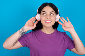 Fototapeta premium Joyful young beautiful tattooed girl wearing blue t-shirt standing against blue background sings song keeps hand near mouth as if microphone listens favorite playlist via headphones