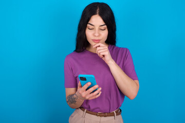 Thoughtful happy young beautiful tattooed girl wearing purple t-shirt standing against blue background hold look phone, copyspace