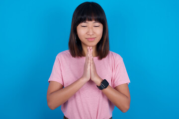 Indoor closeup of young beautiful asian woman wearing pink t-shirt against blue wall practicing yoga and meditation, holding palms together in namaste, looking calm, relaxed and peaceful.