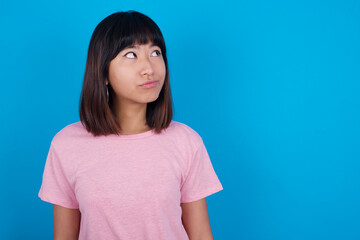 Obraz premium young beautiful asian woman wearing pink t-shirt against blue wall, looks pensively aside, plans actions after university, imagines what to do Thinks over about new project.