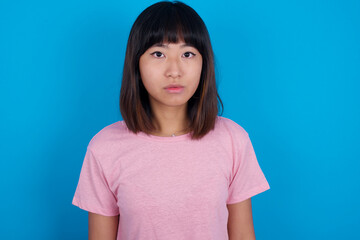 Joyful young beautiful asian woman wearing pink t-shirt against blue wall looking to the camera,...