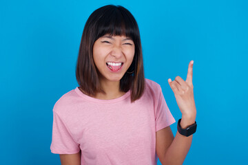 Portrait of a crazy young beautiful asian woman wearing pink t-shirt against blue wall showing tongue horns up gesture, expressing excitement of being on concert of band.