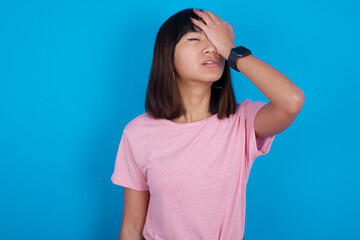 Frustrated young beautiful asian woman wearing purple t-shirt against blue wall holding hand on forehead being depressed regretting what he did having headache, looking stressful.