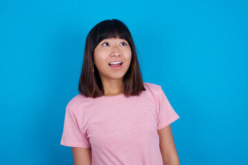 Surprised young beautiful asian woman wearing purple t-shirt against blue wall, shrugs shoulders, looking sideways, being happy and excited. Sudden reactions concept.