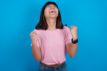 Fototapeta premium Young beautiful asian girl wearing pink t-shirt against blue background looks with excitement up, keeps hands raised, notices something unexpected.