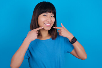 Strong healthy straight white teeth. Close up portrait of happy Young beautiful asian girl wearing blue t-shirt against blue background with beaming smile pointing on perfect clear white teeth.