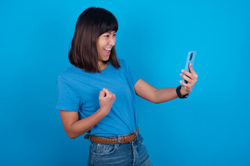 Happy cheerful Young beautiful asian girl wearing blue t-shirt against blue background receiving good news via e-mail and celebrating success while standing and looking at mobile phone.