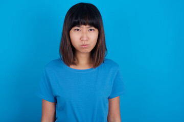Fototapeta premium Offended dissatisfied young beautiful asian girl wearing blue t-shirt against blue background with moody displeased expression at camera being disappointed by something