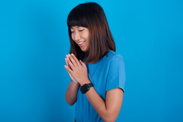 Overjoyed young beautiful asian girl wearing blue t-shirt against blue background laughs joyfully and keeps palms pressed together hears something funny